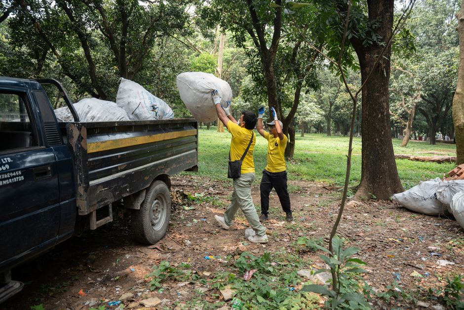 Two workers in yellow shirts are engaged in rubbish removal in a wooded outdoor area, loading large white plastic bags filled with garden waste or debris onto a black flatbed truck. One worker is lifting a bag above his head to place it onto the truck, while the other is preparing to assist, both wearing gloves for safety. The truck is parked on uneven, leaf-covered ground amid trees with green foliage, providing shade and a natural environment. The background shows more trees and scattered debris, indicating a site for clearing garden or green waste. Additional large bags filled with waste are visible on the ground nearby. The scene portrays an outdoor waste clearance activity, reflecting private or independent rubbish collection efforts, supporting services like rubbish removal offered by companies such as Rubbish Removal Watford. Natural lighting highlights the textures of the bags' plastic material and the earthy surroundings, presenting a clear, factual depiction of onsite waste removal in a park or garden setting.