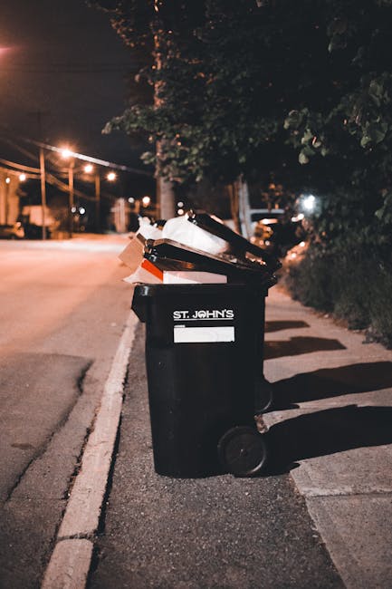 A black plastic waste bin labeled 'St. John's' is situated on a sidewalk beside a curb in an urban residential area at night. The bin's lid is open, revealing a mixture of domestic rubbish, including paper, cardboard, and plastic items. The bin appears to be made of sturdy textured plastic with large wheels at the rear, and there are a few discarded items leaning against the side. The surrounding environment includes dim street lighting casting elongated shadows and a background of mature trees and shrubbery, with a few lights from nearby houses or buildings visible at a distance. The scene suggests an area serviced by private waste collection, with the bin positioned for rubbish removal services related to domestic waste management, aligned with the context of independent or alternative waste disposal methods often used in private rubbish removal operations in Watford.