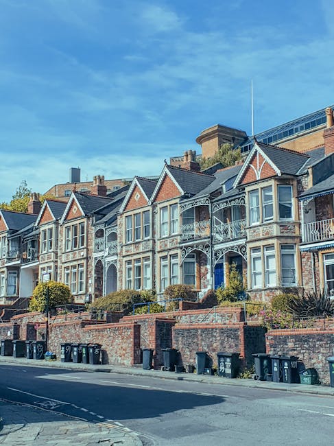 A narrow residential street lined with red-brick terraced houses featuring pitched slate roofs, chimneys, and small front yards or steps leading up to doors. On the right side, a tall wooden fence runs parallel to the pavement, partially obscuring a garden with dense, dark green foliage. The pavement appears worn and uneven, with a slight curve in the street. Several black wheelie bins are visible along the curb, suggesting private waste collection, while the scene is illuminated by soft natural light, casting shadows on the ground. In the background, additional houses extend into the distance under a partly cloudy sky, creating a typical suburban setting that could relate to independent rubbish removal or on-site clearance activities managed by companies like Rubbish Removal Watford.