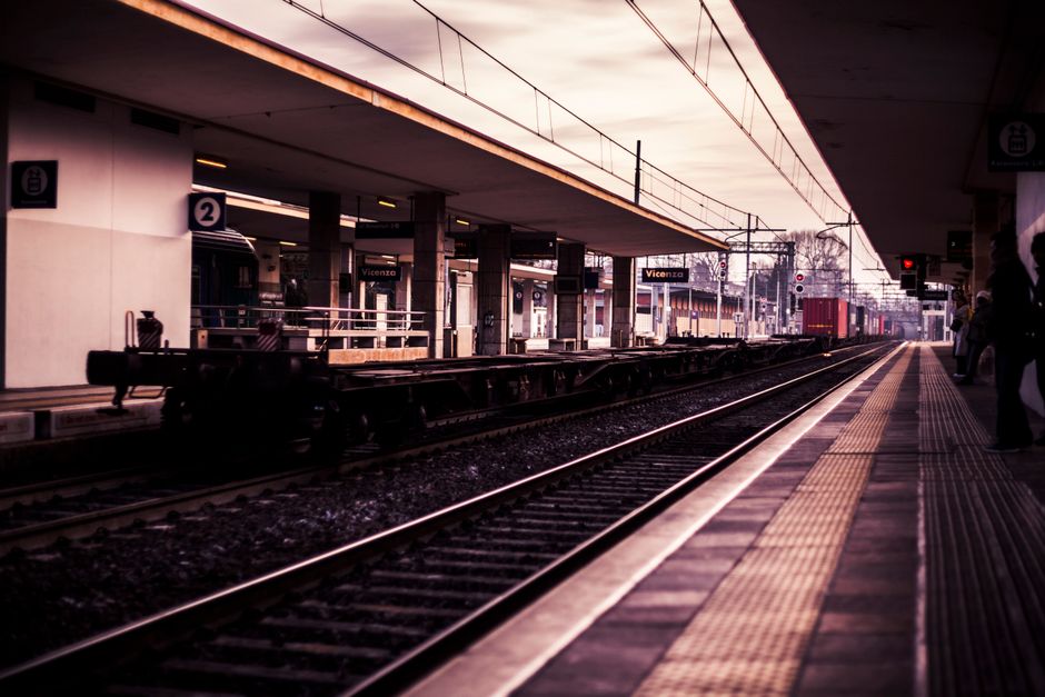 A view of a train station platform under an elevated railway track, with several dark-colored trains and freight cars visible on the tracks in the background. The platform has tactile paving strips running parallel to the tracks, which are yellowish-brown with textured surface for safety. On the left side of the image, a small area of the station building can be seen, displaying signs with the station name 'Vicenza' attached to concrete pillars supporting the upper structure. The station's roof extends over the platform, with a smooth, dark underside, and faint lighting fixtures are mounted along the ceiling. In the distance, there are a few passengers waiting, some standing near the edge or leaning against the pillars, dressed in winter clothing. Overhead electrical wires run above the railway lines, supported by tall metal poles, contributing to the overall urban railway setting. The natural lighting suggests late afternoon or early evening, casting a pinkish hue over the scene, with a slightly overcast sky in the background.