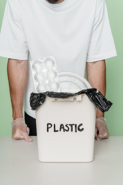 A person wearing a white t-shirt and transparent gloves is positioned behind a white plastic bin labeled 'PLASTIC', which contains a black trash bag and a clear plastic egg carton with six circular depressions. The bin is set on a flat surface in front of a light green background, suggesting an indoor environment focused on waste segregation or recycling. The egg carton is partially visible, leaning against the inner rim of the bin, with the black trash bag loosely draped over it, indicating an organized approach to rubbish disposal. The scene reflects a conscientious effort towards separating plastic waste for collection or recycling, aligning with private rubbish removal services that support proper waste handling and environmentally responsible disposal practices, particularly for recyclable materials like plastics. This setup underscores the importance of efficient waste segregation in the context of rubbish collection and disposal solutions offered by companies such as Rubbish Removal Watford, emphasizing accurate sorting and environmentally conscious waste management.