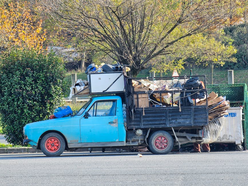 A small, old blue pickup truck with a black cargo bed is parked on a street beside a curb, filled with a large amount of mixed waste materials including flattened cardboard boxes, black plastic bags, and other discarded items. The truck's wheels are painted red, and its body shows signs of wear with some scratches and faded paint. The cargo area is loaded high with rubbish, some of which appears loosely stacked and secured with ropes. Behind the truck, there is a green waste collection container, possibly used for disposal or recycling, positioned on the pavement. In the background, several mature trees with green and autumn-colored leaves are visible, indicating a park or residential area setting. The scene is illuminated by natural daylight, creating a clear, detailed view of the cluttered vehicle that could be part of a private waste collection or rubbish removal service, such as Rubbish Removal Watford, engaged in on-site clearance or alternative waste handling outside of local authority rubbish collection services.